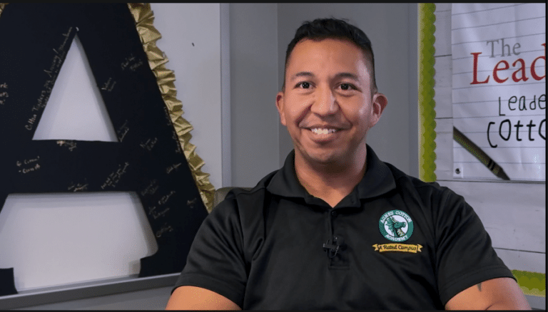 A man in a black polo shirt with a circular logo sits smiling in an office, embodying the spirit of a Certified Champion Educator. Behind him, the large letter "A" and motivational posters from VentureLab set the stage for empowering young minds.